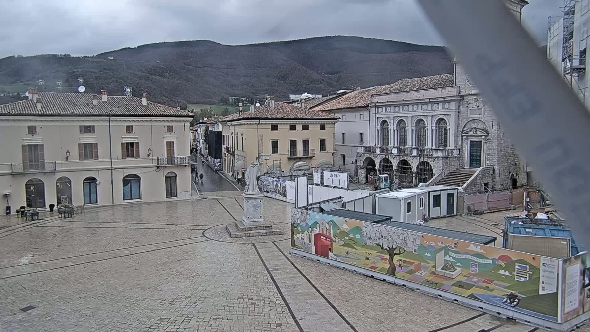 A town square with several buildings, a statue, and construction barriers under a cloudy sky.