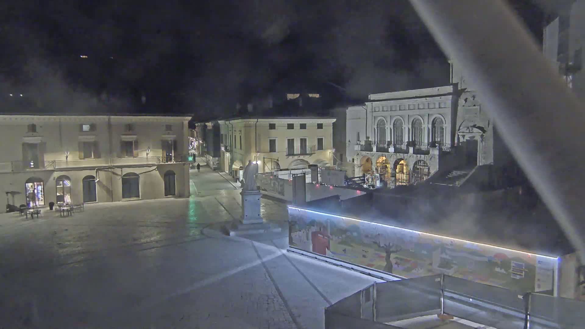 A nighttime view of an empty town square with several buildings, a statue, and a large structure with artwork displayed on its side, under hazy or foggy conditions.