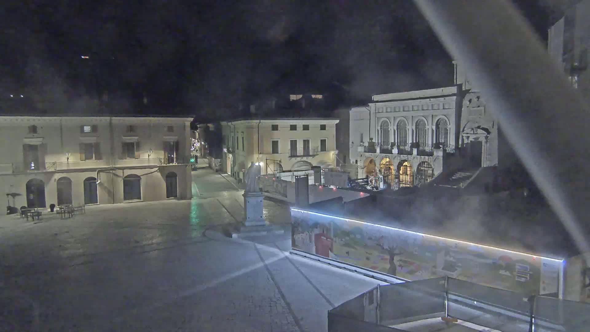 A nighttime view of a town square with several buildings, a statue, and a construction site featuring a painted wall.