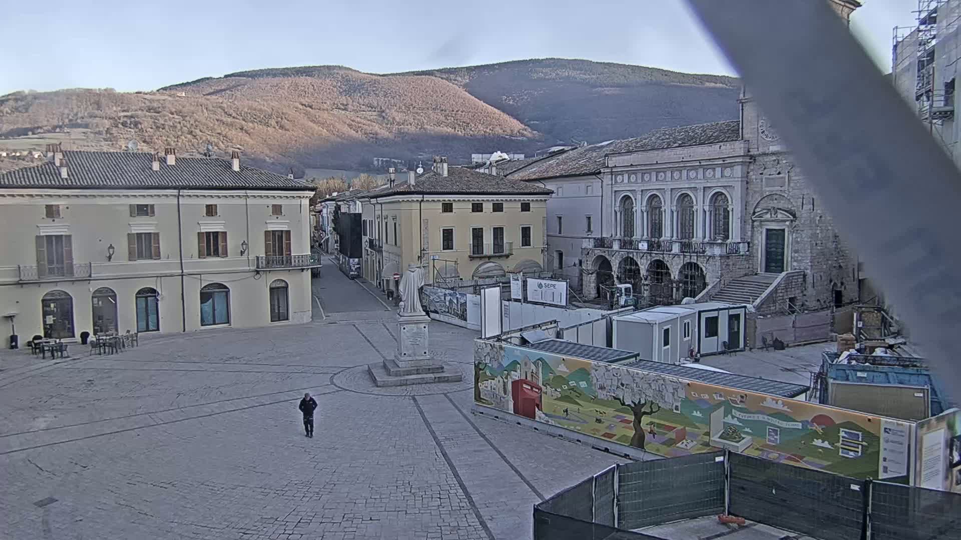 A mostly empty town square with several buildings, construction materials, and a statue, under a clear sky, is set against a backdrop of distant mountains.