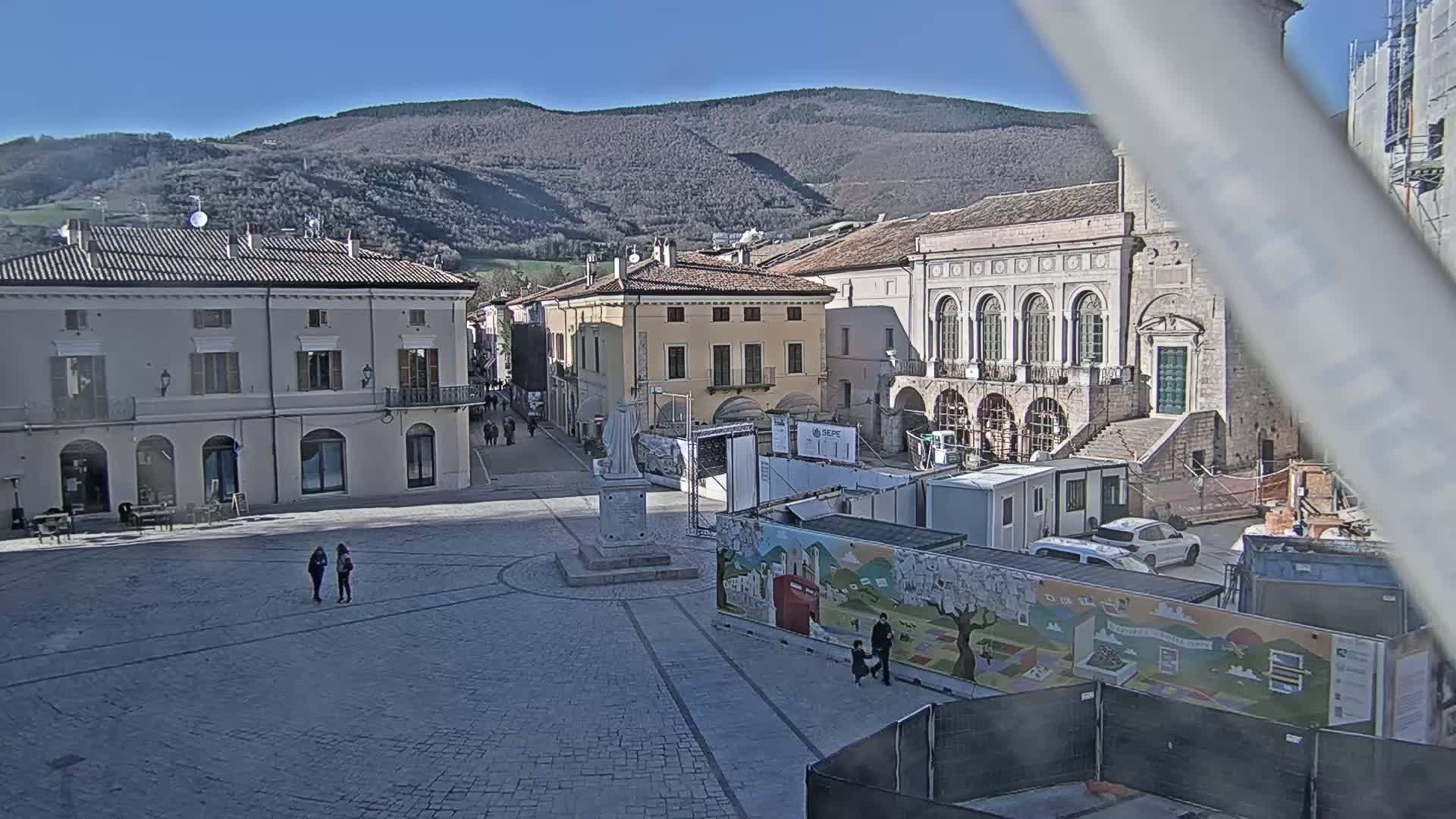 A town square with several buildings, a monument, and construction materials is visible under a sunny sky with mountains in the background.