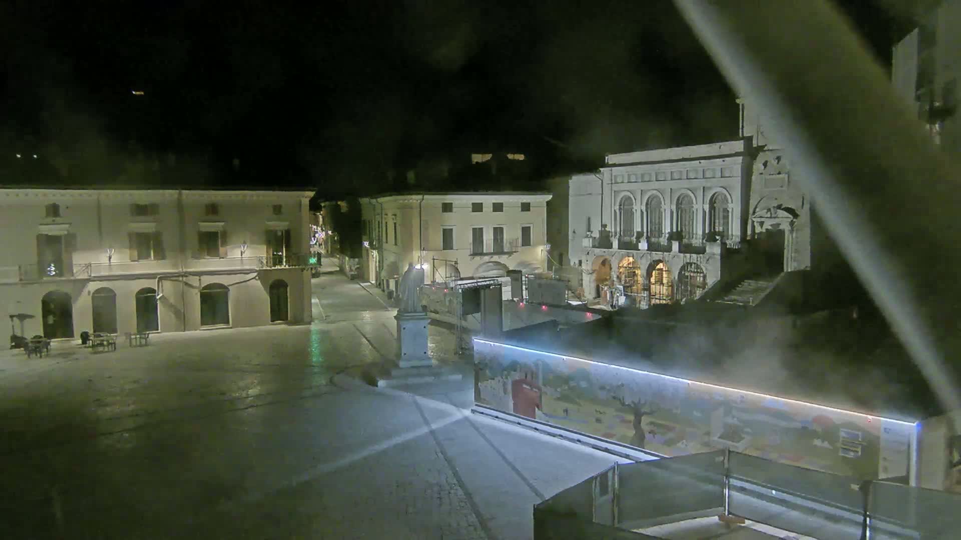 A nighttime view of a town square with several buildings, a statue, and what appears to be a temporary structure with a painted mural, under hazy or foggy conditions.