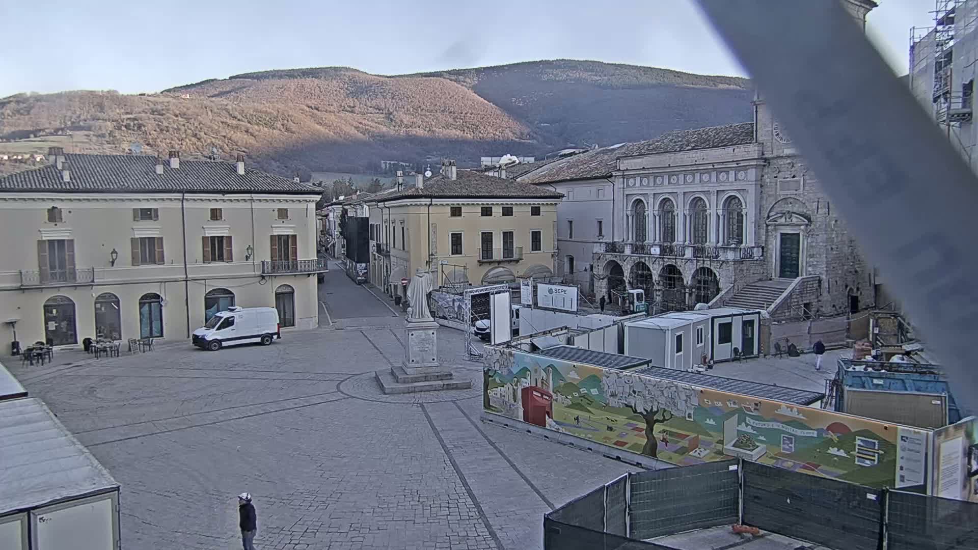 A town square with several buildings, a statue, and construction barriers, situated against a backdrop of mountains under a clear, bright sky.