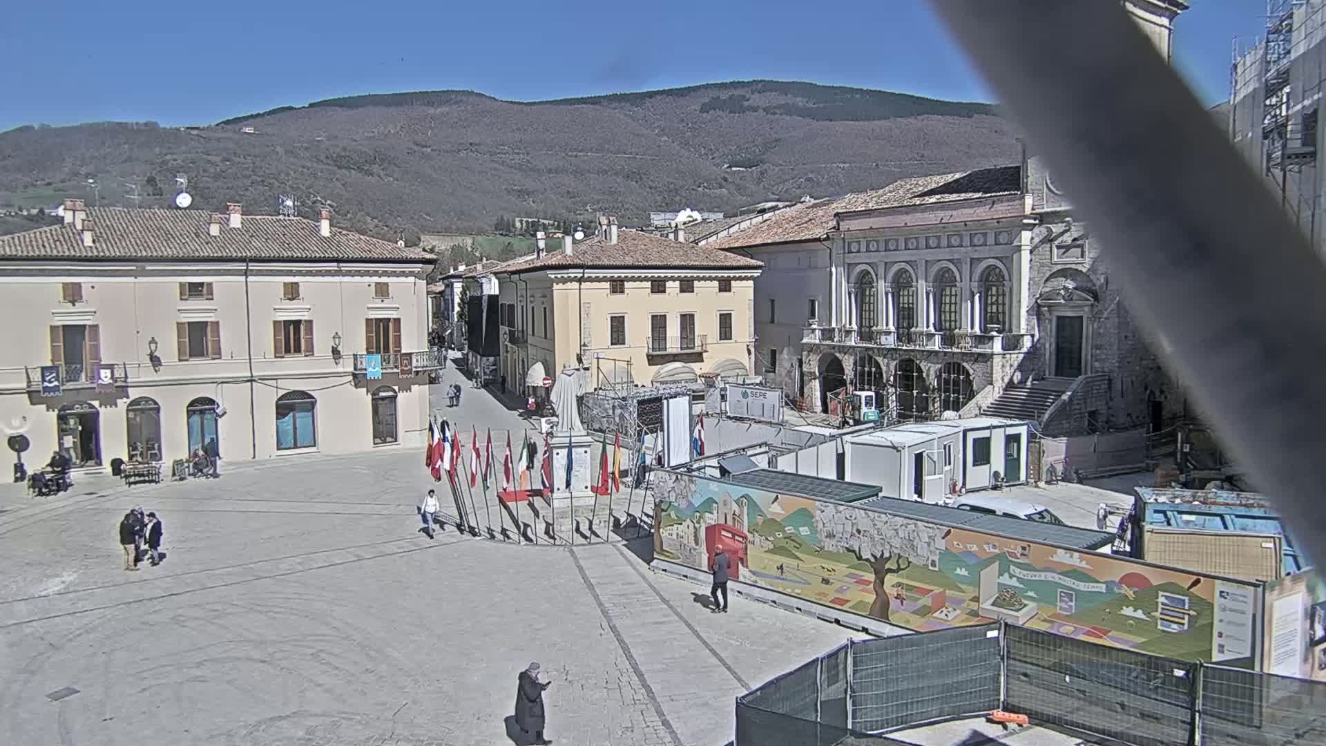 A town square with several buildings, a monument, and various temporary structures is visible under a sunny sky, with mountains in the background.