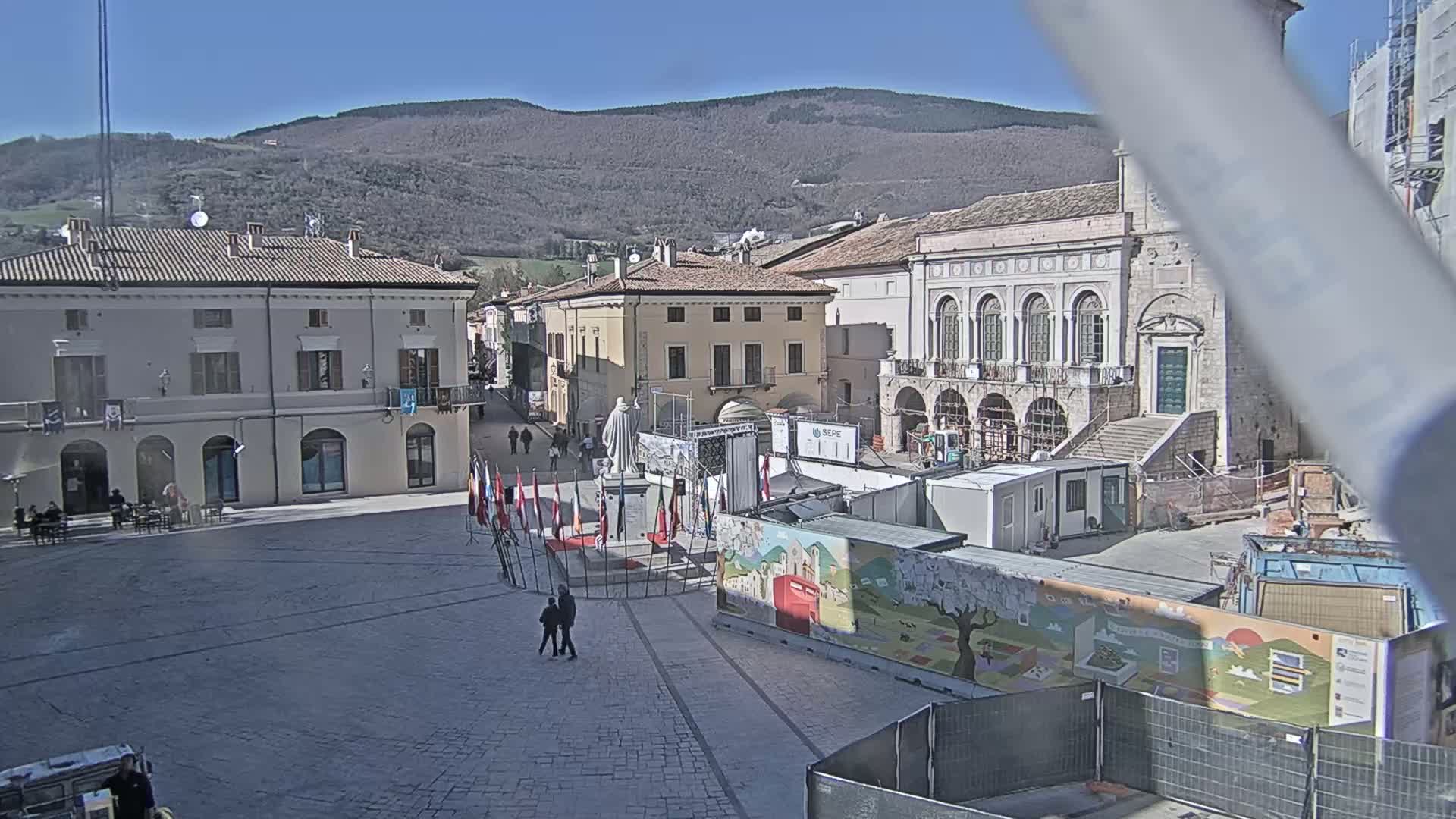 A town square with several buildings, a statue, and construction materials is visible on a sunny day with mountains in the background.