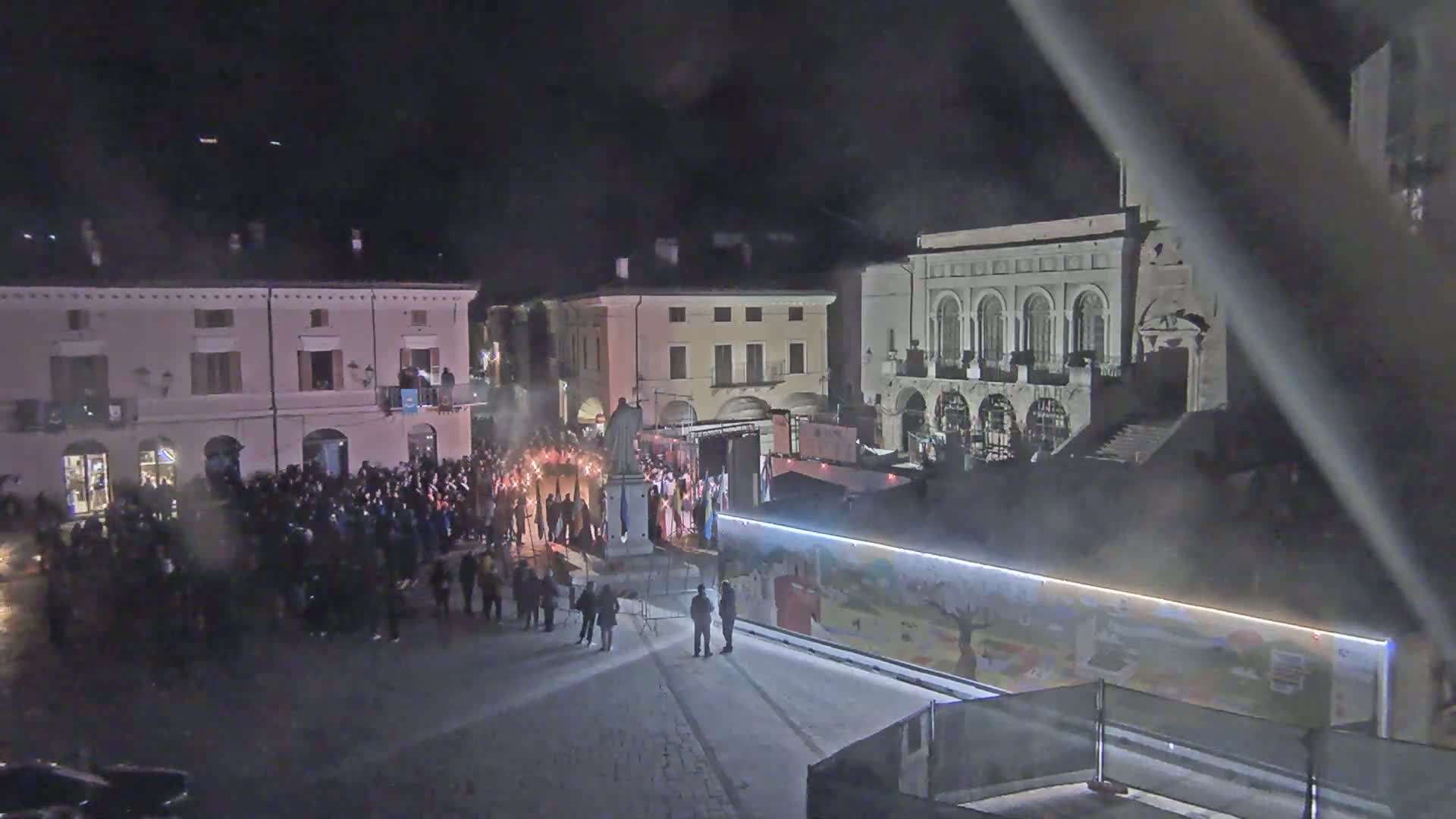 A nighttime outdoor scene shows a large crowd gathered in a town square, some holding sparklers, with buildings in the background.