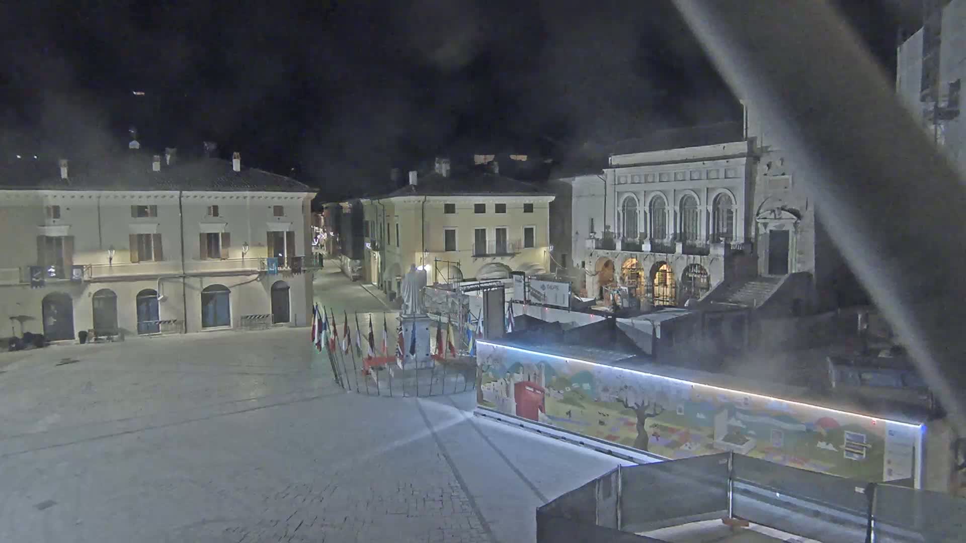 A nighttime view of a town square with several buildings surrounding it, a statue in the center, and a decorated container in the foreground.