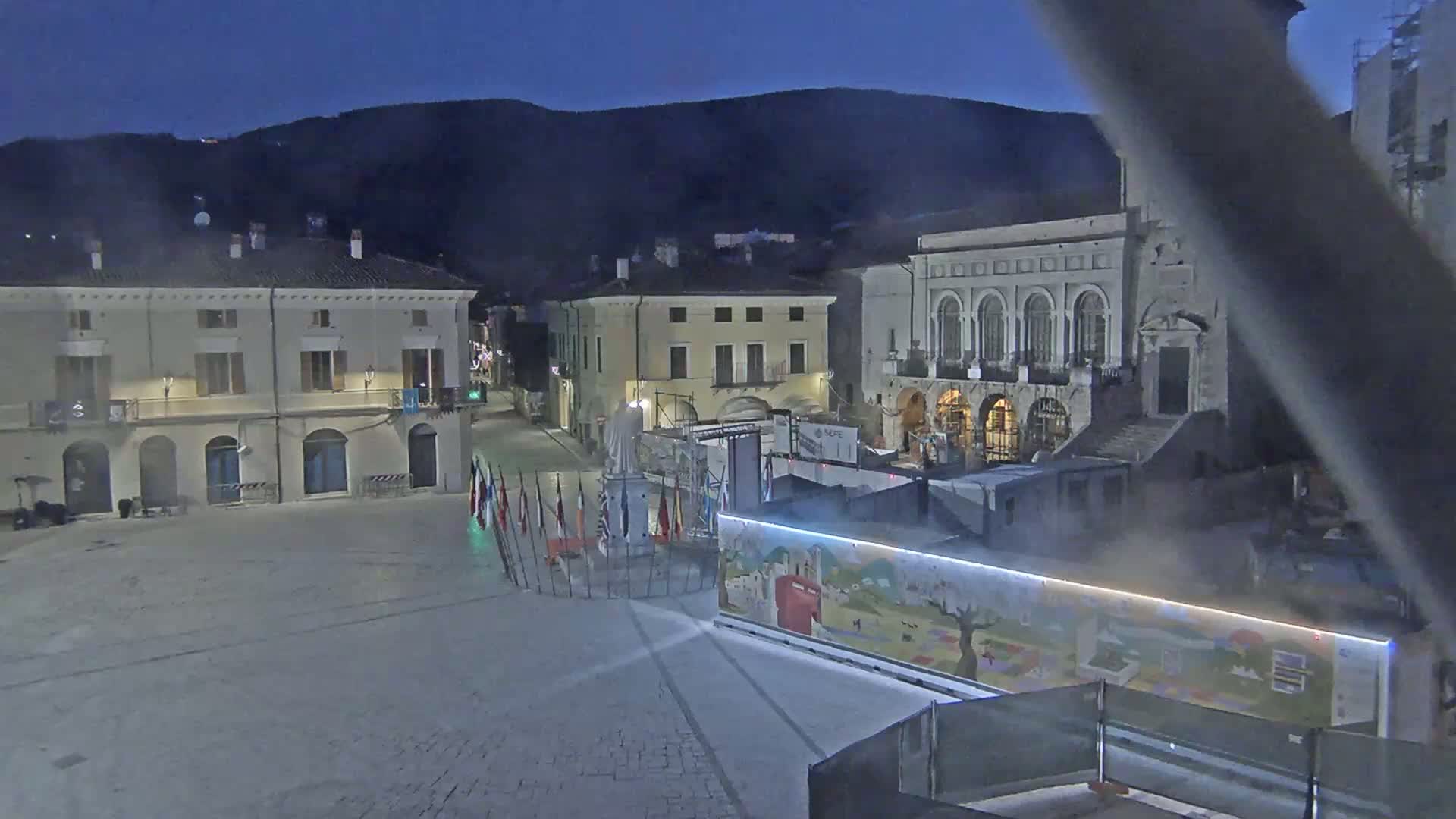 A town square at night, under a dark sky, features several buildings, a statue, and numerous flags displayed near a colorful, illustrated structure.