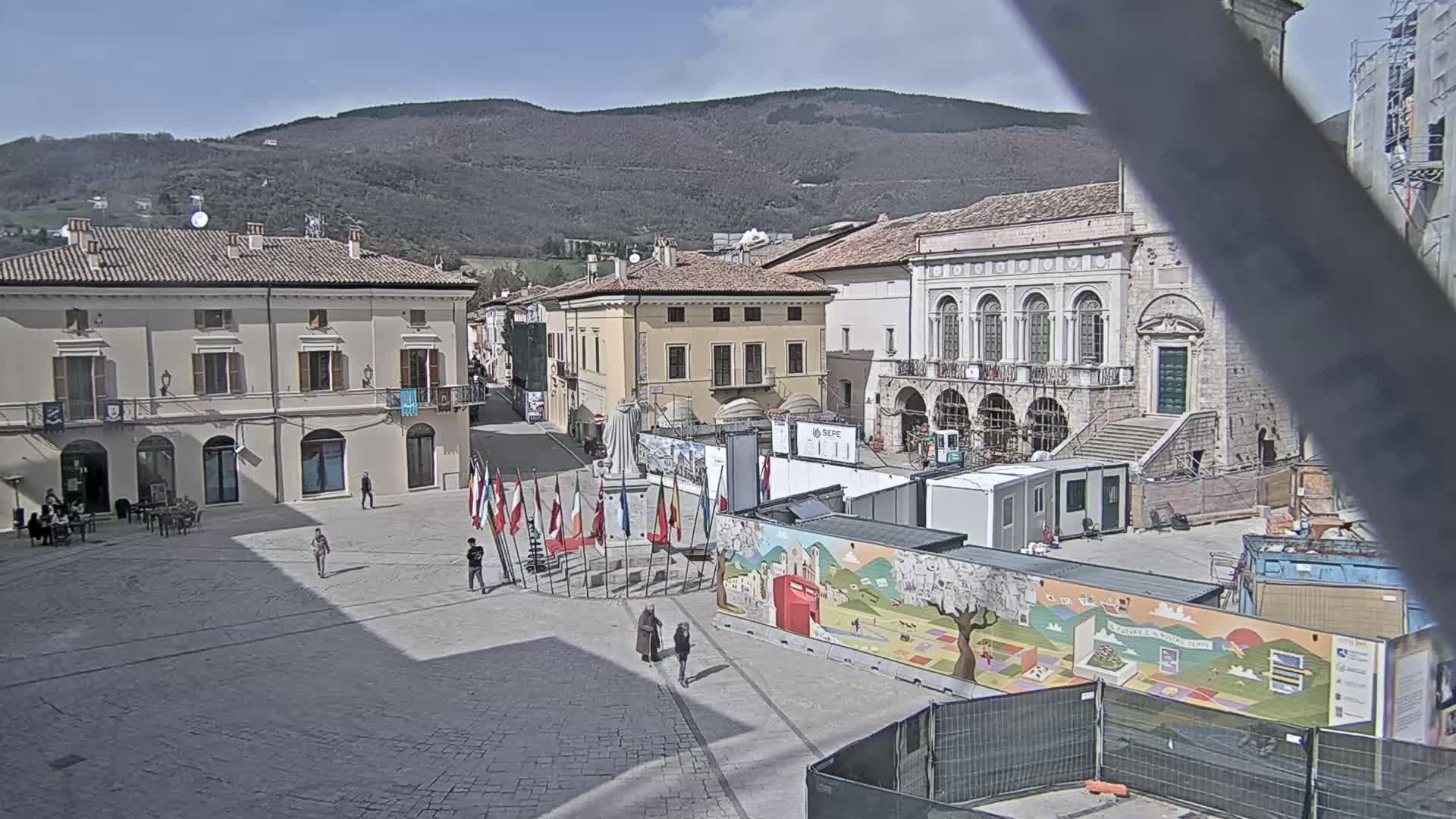 A sunny town square with several buildings, construction materials, and flags, situated in front of a mountain range.