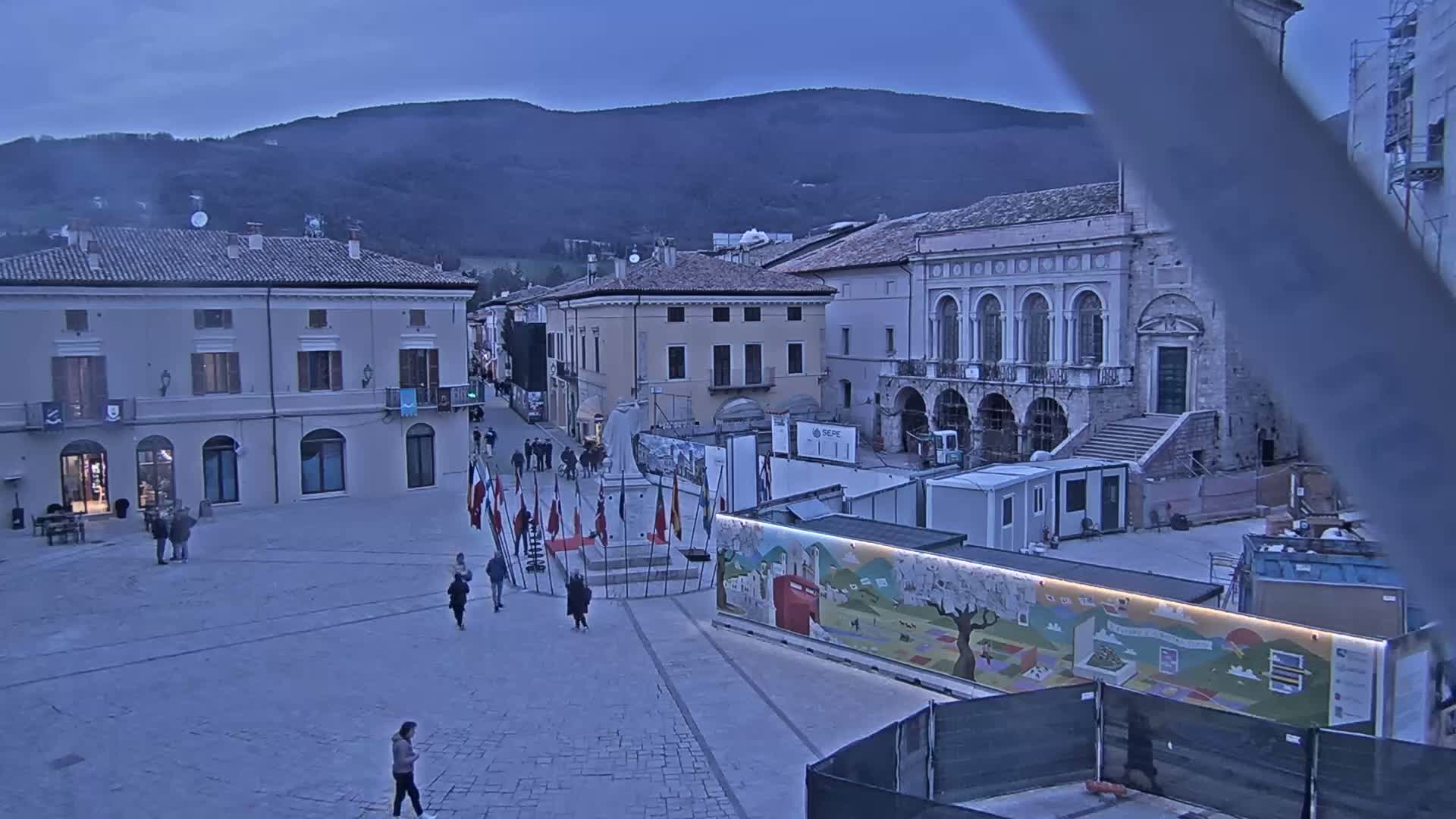 A town square, under a twilight sky, features several buildings, construction equipment, and a few pedestrians;  mountains are visible in the background.