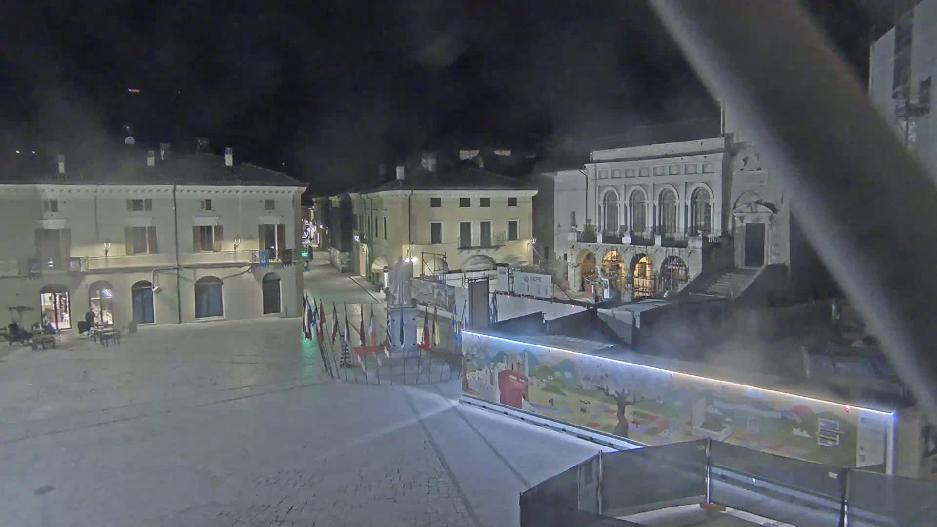 A mostly empty town square at night, under hazy conditions, features several buildings, a statue, and a long structure with artwork painted on its side.