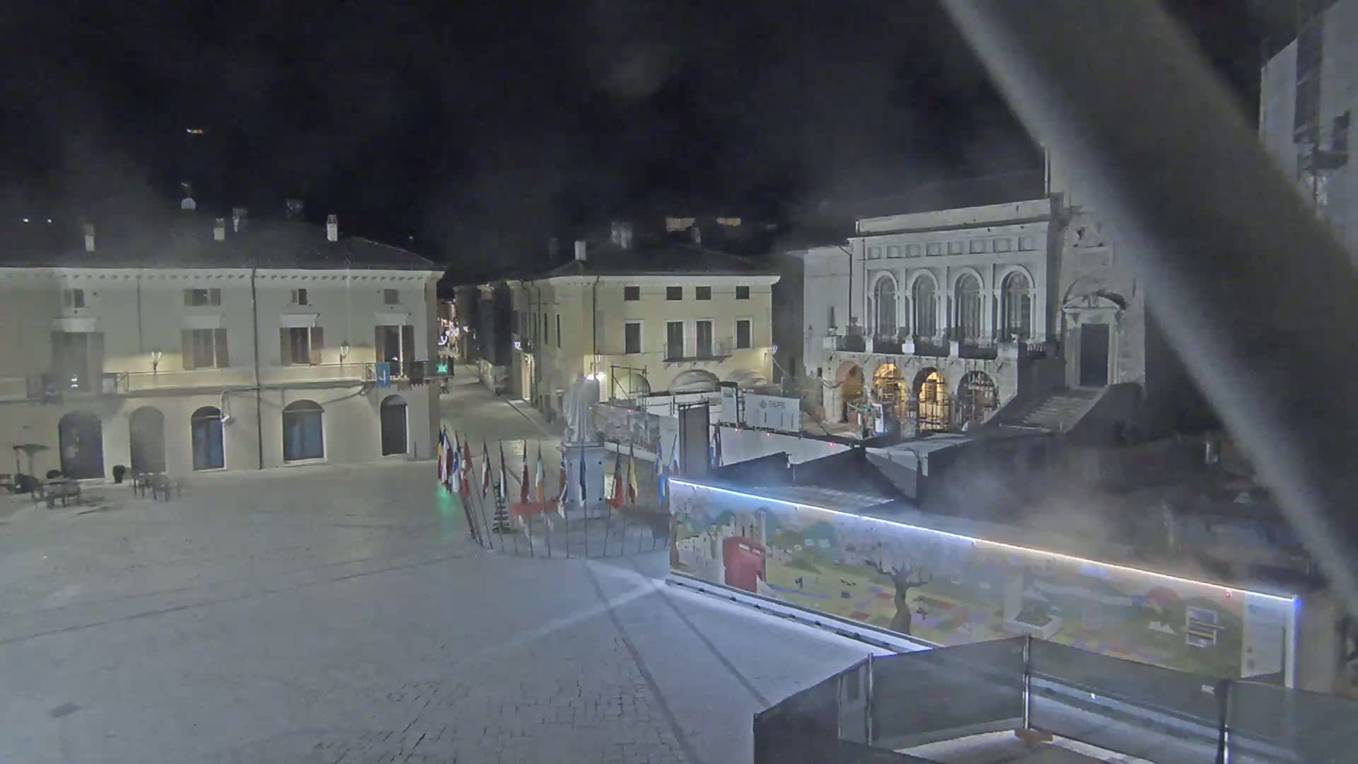 A nighttime view of a town square with several buildings, a statue, and flags, under hazy or foggy conditions.