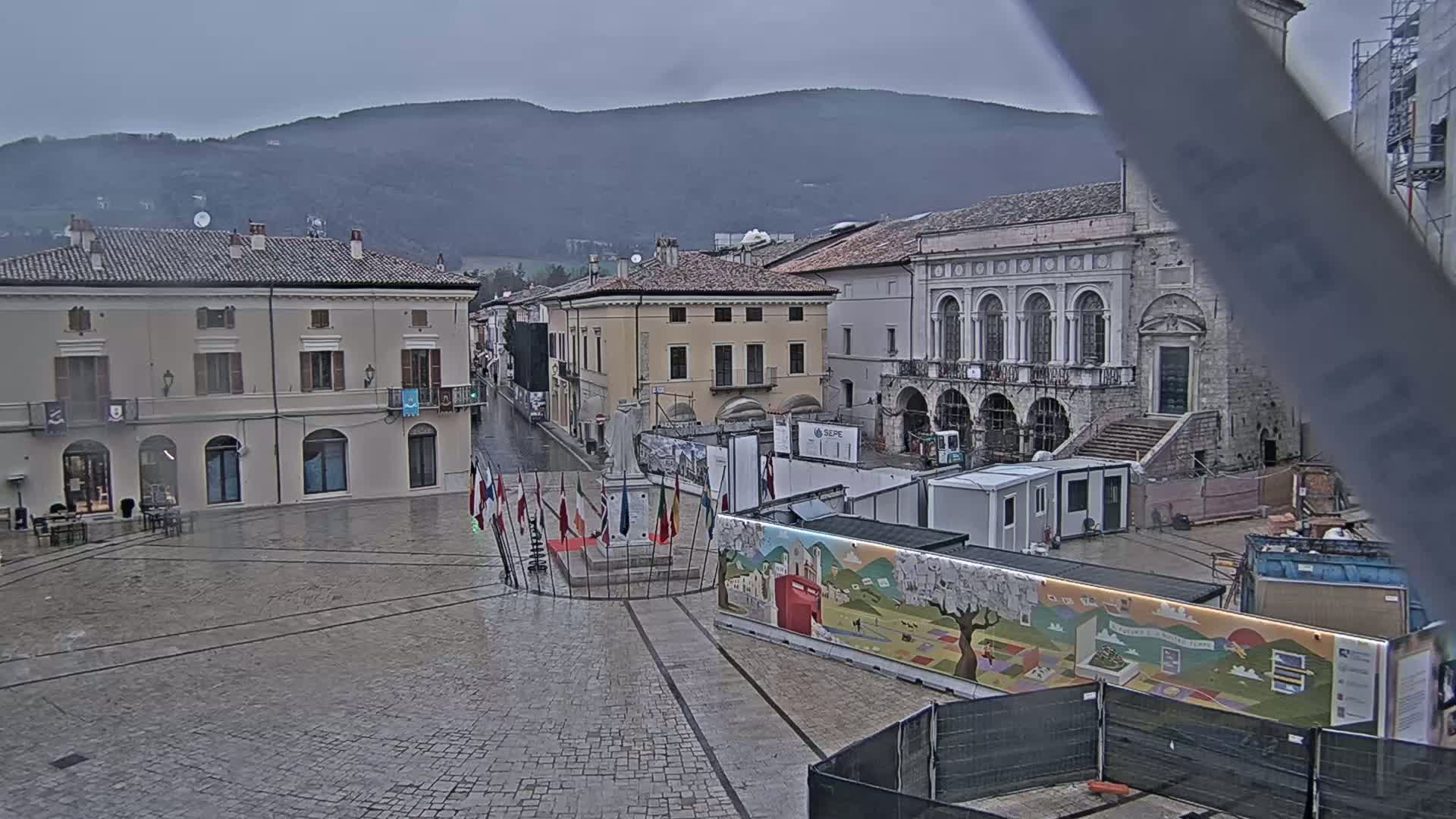 A town square with several buildings, a monument, and construction barriers under an overcast sky.