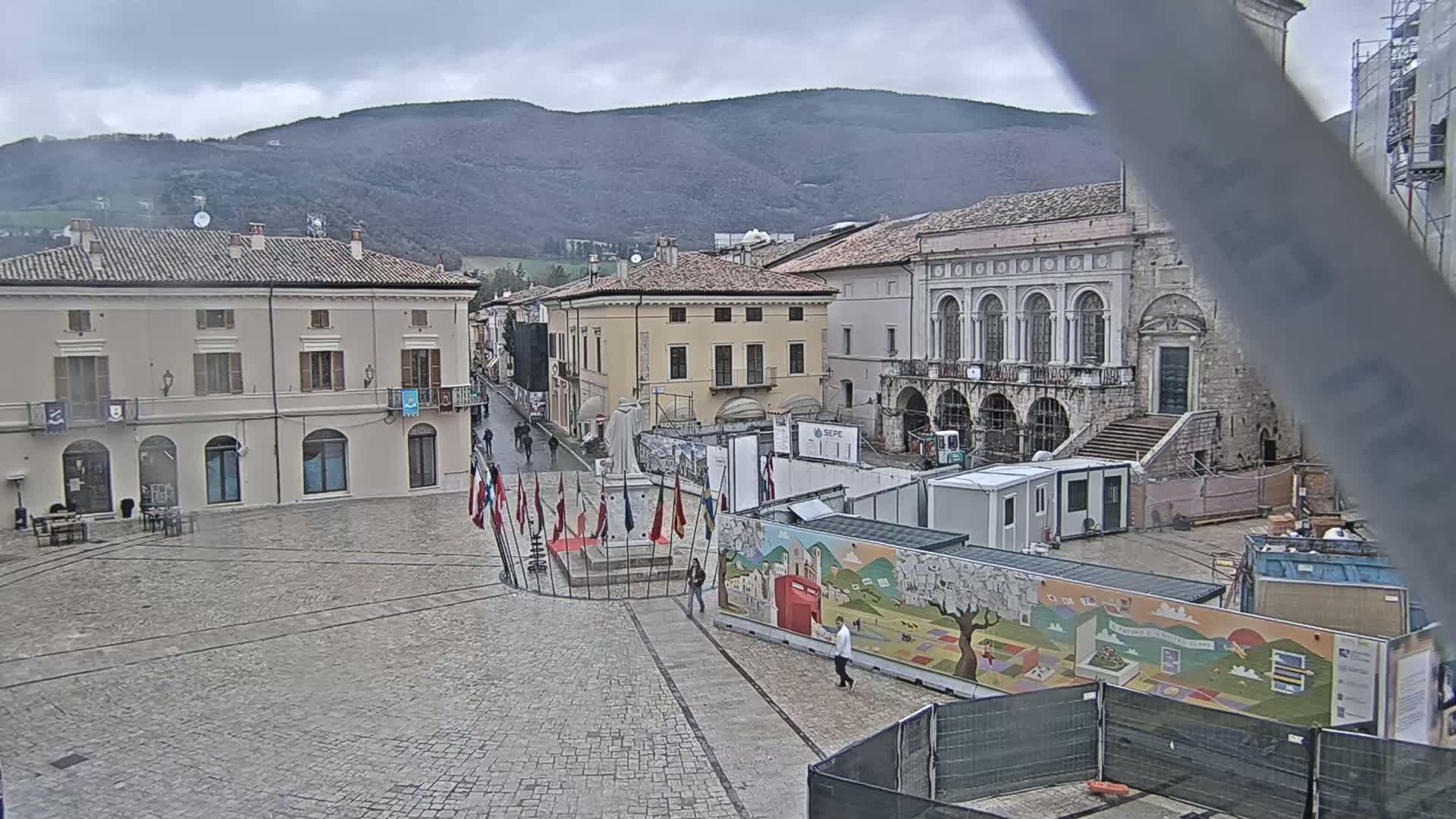 A town square with several buildings, a statue, and construction barriers is seen under an overcast sky.