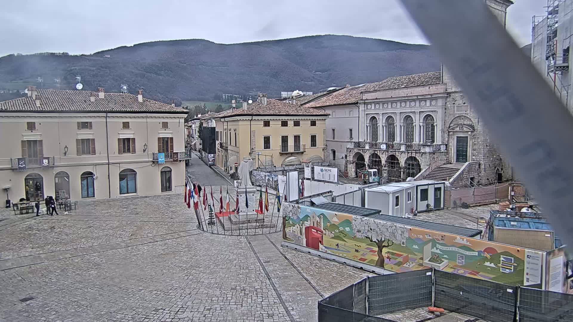 A town square with several buildings, under a cloudy sky, features a monument surrounded by flags and construction barriers.