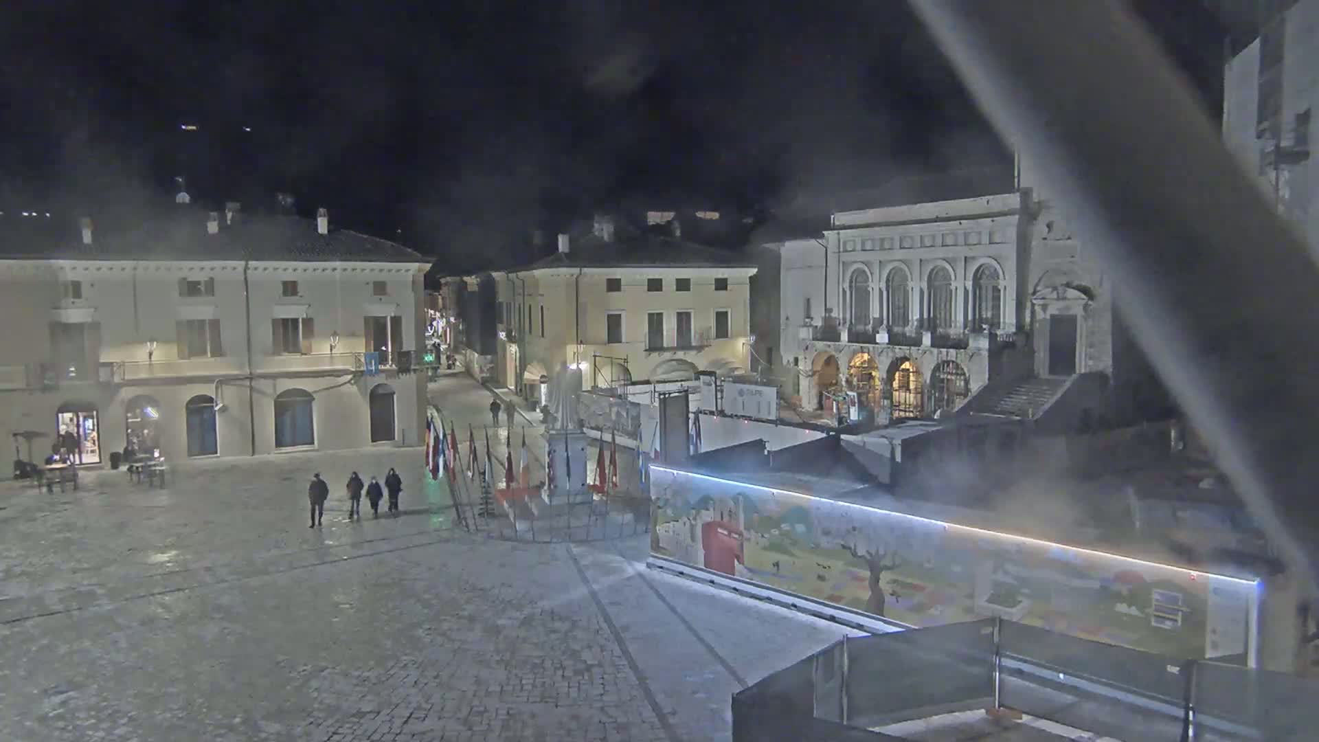A mostly empty town square at night, with several buildings surrounding it, under a hazy sky.