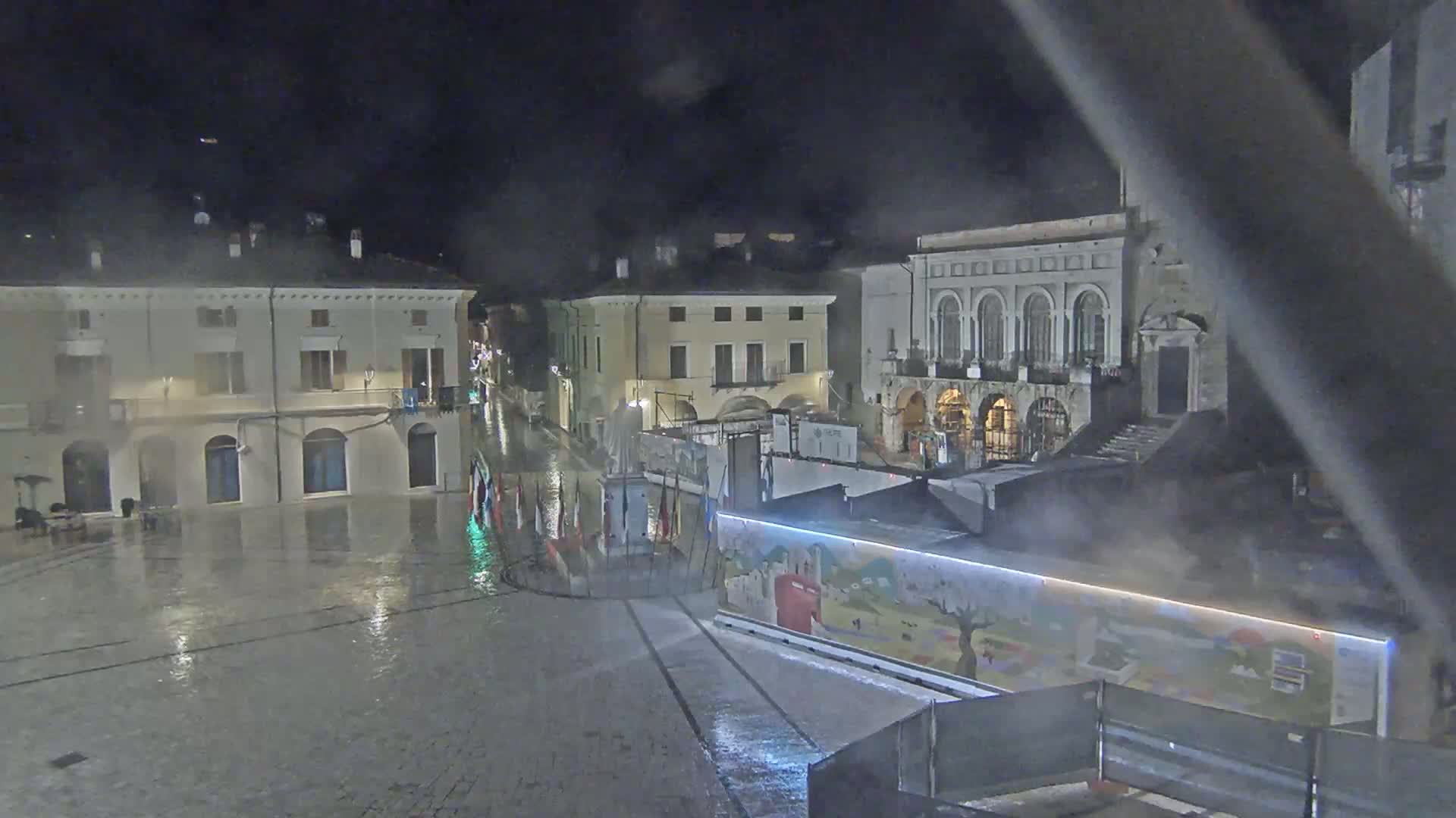 A rain-slicked town square at night, surrounded by historic buildings, features temporary structures and flags in the foreground.