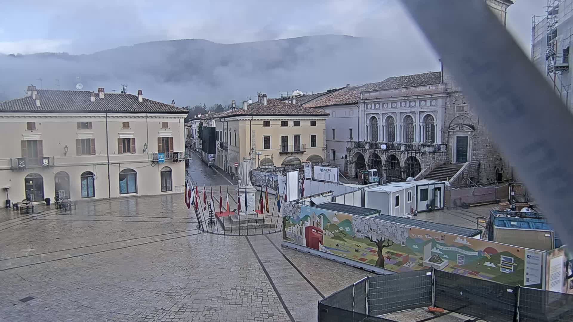 A wet town square with several buildings, a monument surrounded by flags, and construction barriers, all under an overcast sky with mountains in the background.