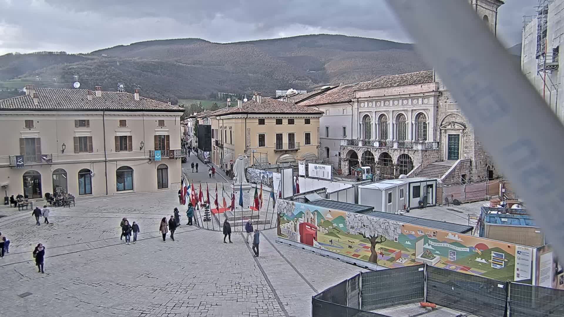 A town square with several buildings, some under construction, is shown under an overcast sky; people walk in the square, and various flags are displayed near a monument.