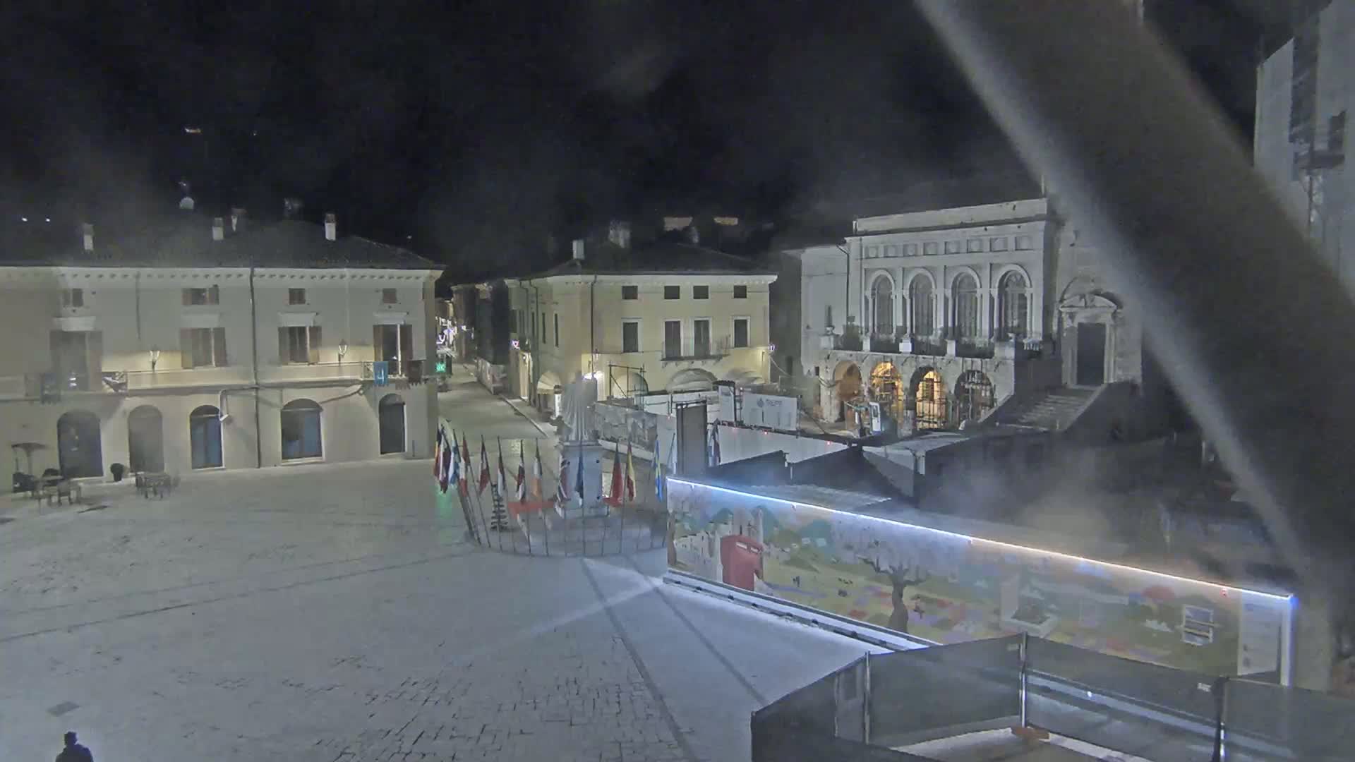 A nighttime view of a snow-covered town square with several buildings, a statue, and a colorful structure with a mural.