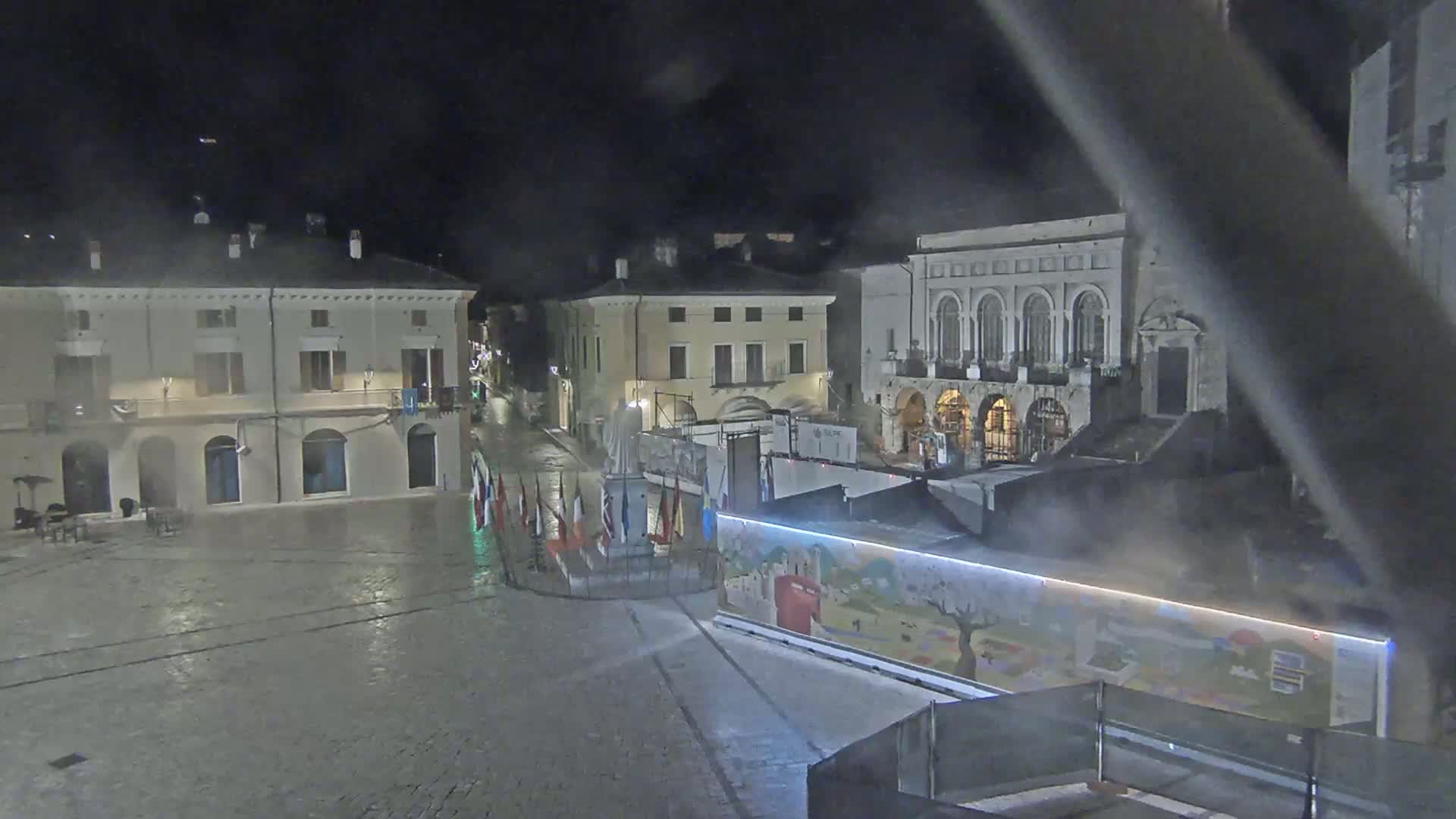 A nighttime view of a town square with several buildings, a statue, flags, and a large illustrated structure, under misty or foggy conditions.