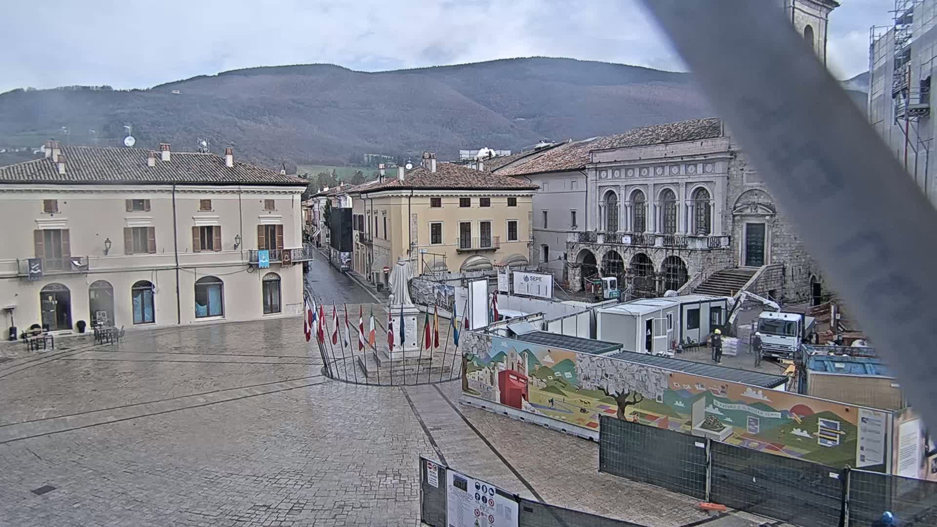A town square with several buildings, a statue, and construction materials is visible under an overcast sky, with mountains in the background.
