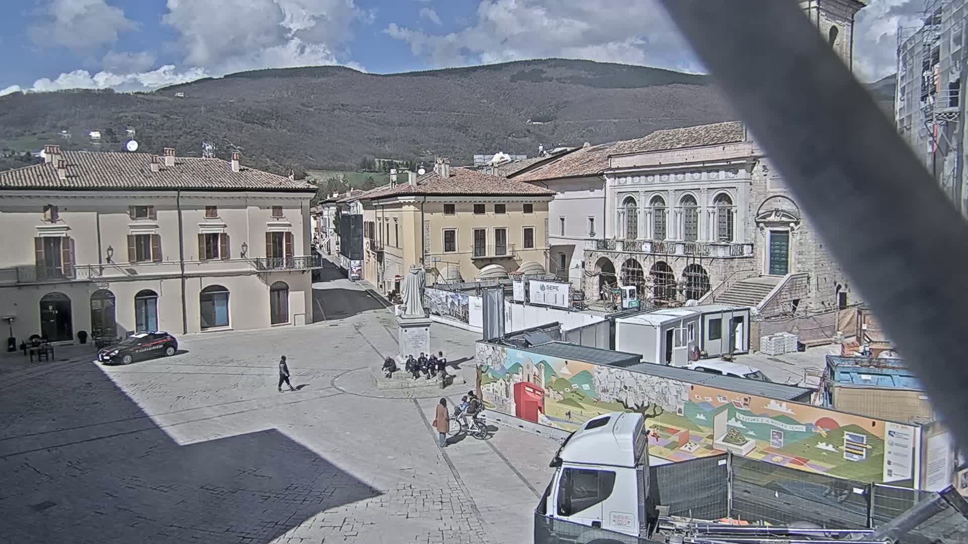 A mostly sunny town square with several buildings, a statue, a few people, construction equipment, and a mountain in the background.
