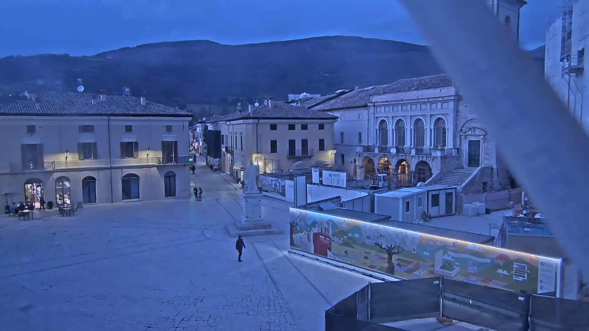 A twilight view of a town square with several buildings, a statue, and construction materials, under a dark blue, clear sky.