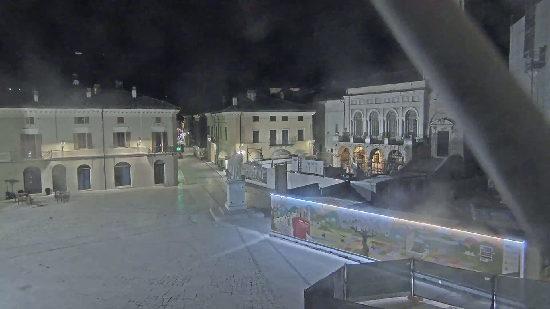 A mostly empty town square at night, illuminated by ambient light, features several buildings and a statue on a pedestal, with a brightly colored mural-covered structure in the foreground.