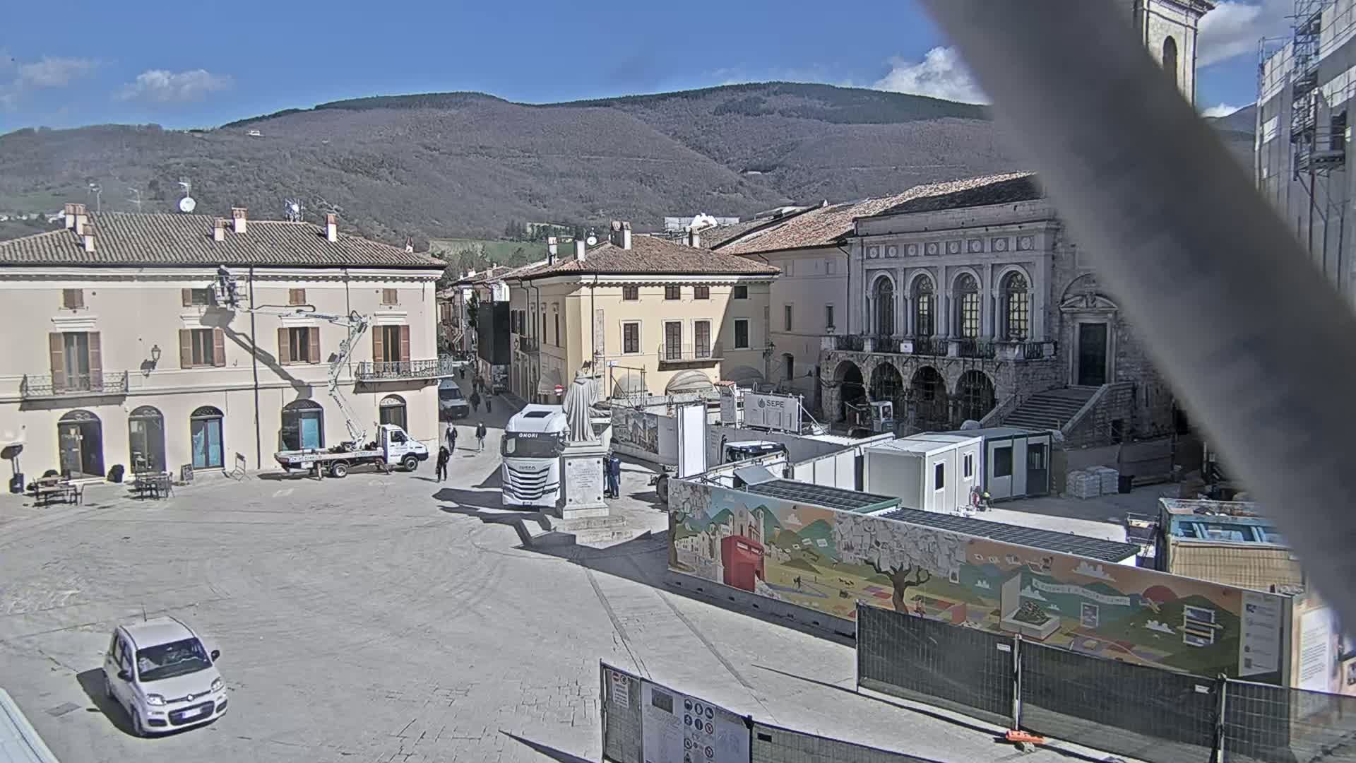 A town square under a sunny sky is partially under construction, with several vehicles and construction materials present, and mountains visible in the background.