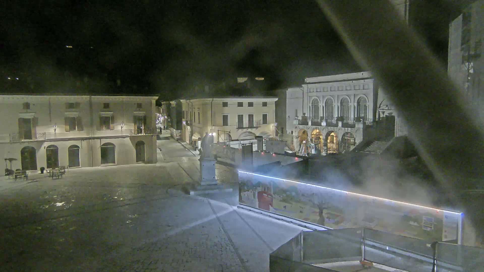 A nighttime view of a town square with several buildings, a statue, and a large construction-style barrier with a mural, under foggy conditions.