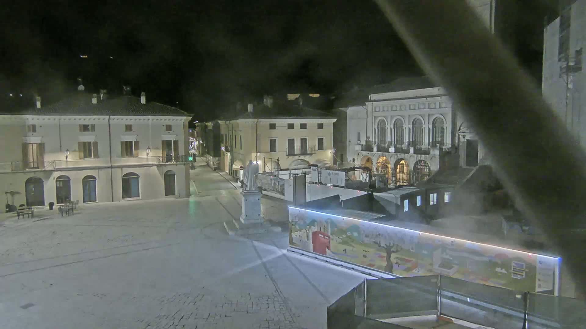 A snow-covered town square at night is illuminated, with several buildings visible, including one with arched windows, and construction barriers with colorful artwork.