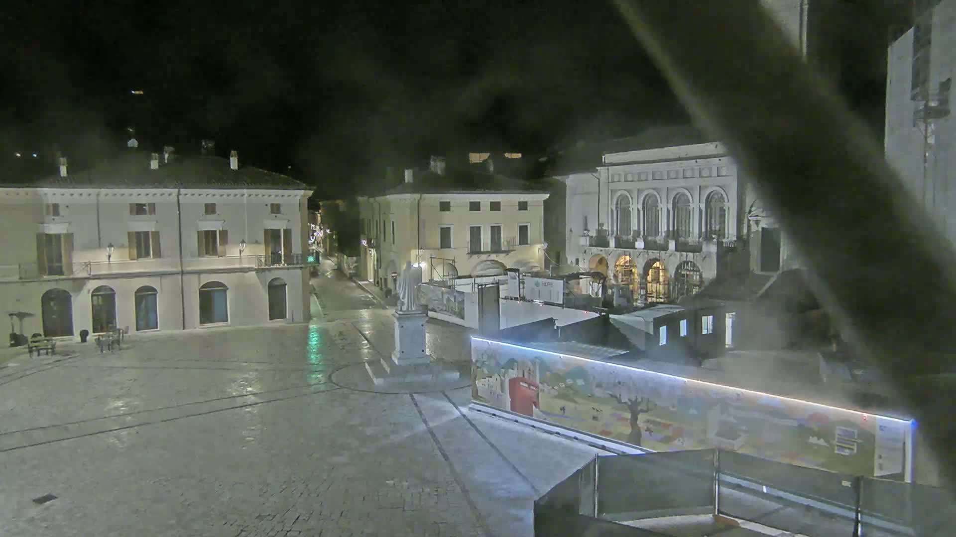 A nighttime view of a town square with several buildings, a statue, and temporary structures, illuminated under hazy conditions.