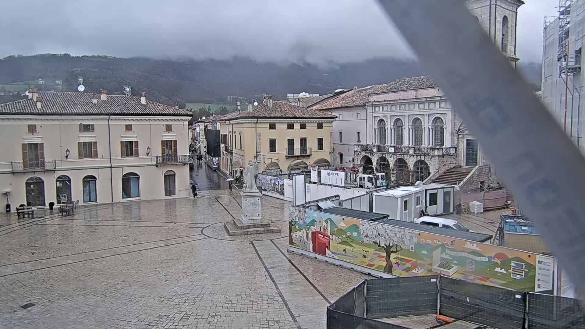 A rain-soaked town square, surrounded by historic buildings and construction, sits under a cloudy sky with mountains in the background.