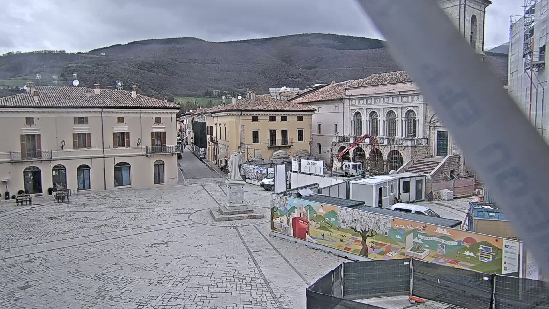 A cobblestone town square, under an overcast sky, is partially obstructed by construction equipment and temporary fencing featuring a colorful mural.