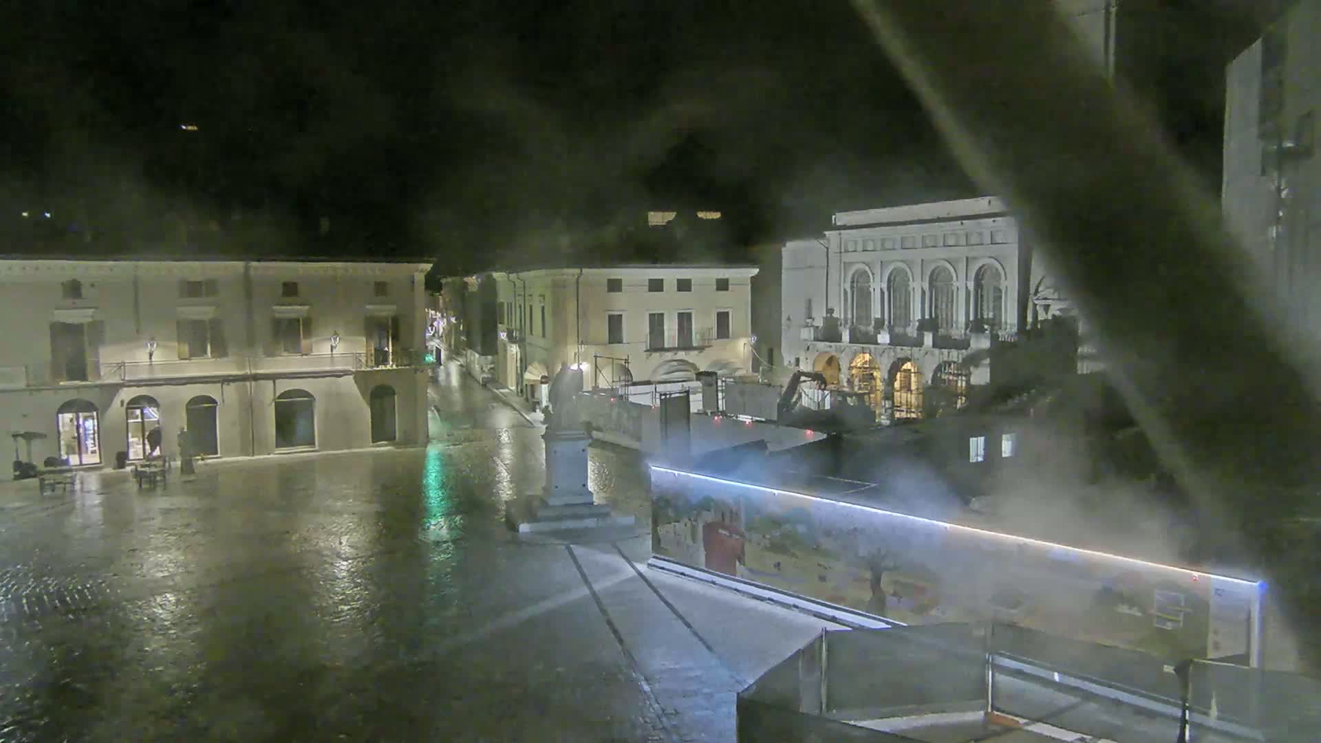 A rain-slicked town square at night, featuring several buildings and a monument, with construction equipment and barriers visible.