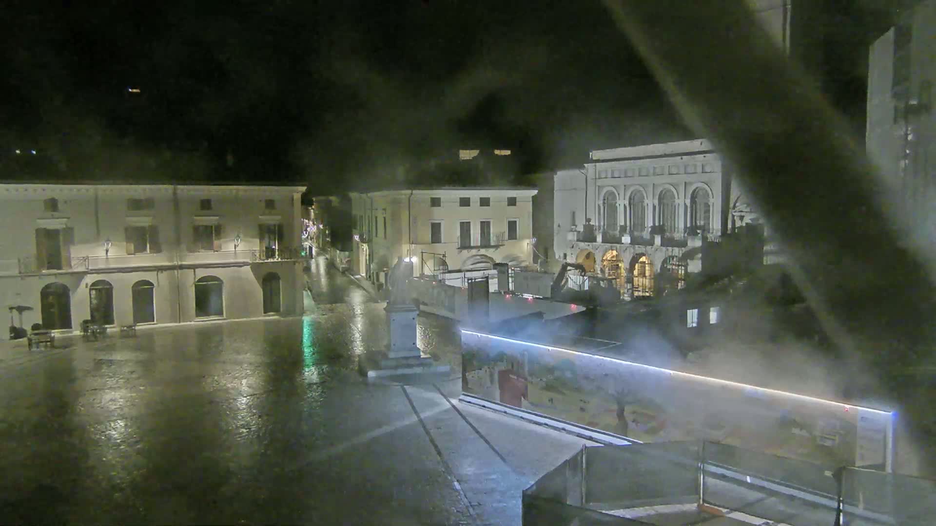 A rain-soaked town square at night is seen, featuring several buildings, a statue, and construction barriers.