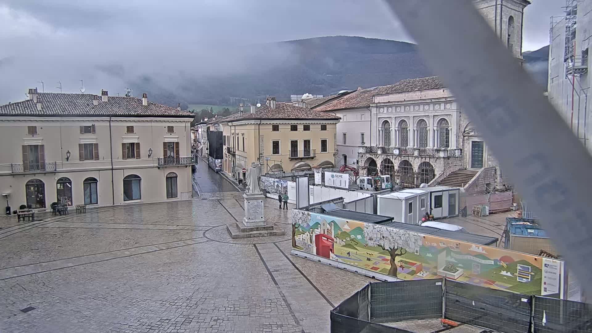 A town square, under an overcast sky, features several buildings, construction materials, and a statue in the center.