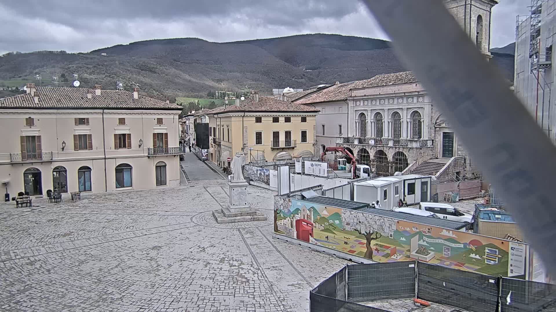 A large, stone plaza in a town nestled against mountains under an overcast sky features construction underway, including temporary structures and a mural.
