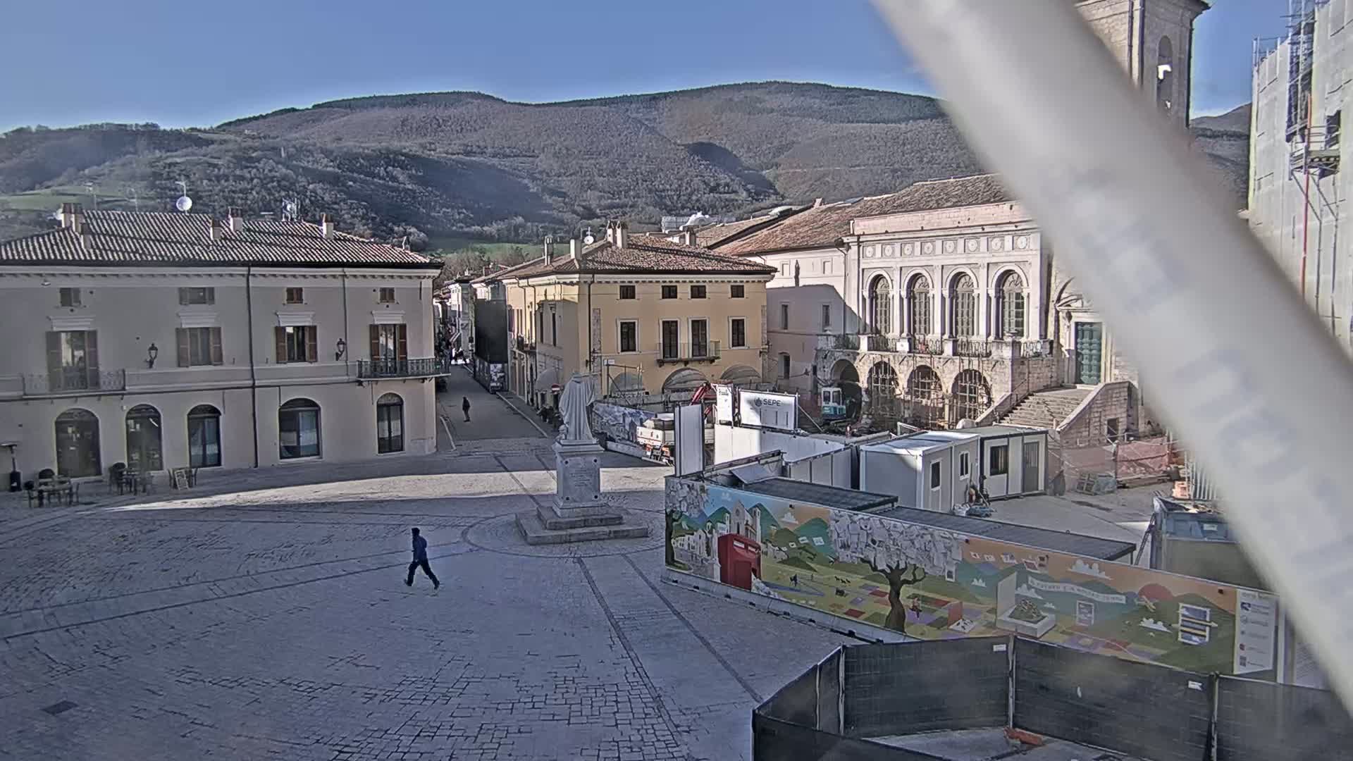 A town square with several buildings, a statue, and construction equipment is viewed on a sunny day with mountains in the background.