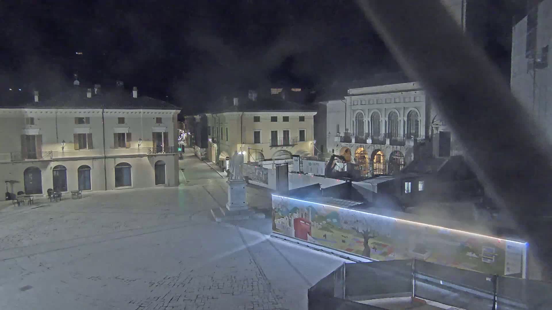 A snow-covered town square at night is surrounded by buildings, one of which is under construction, with a statue in the center.