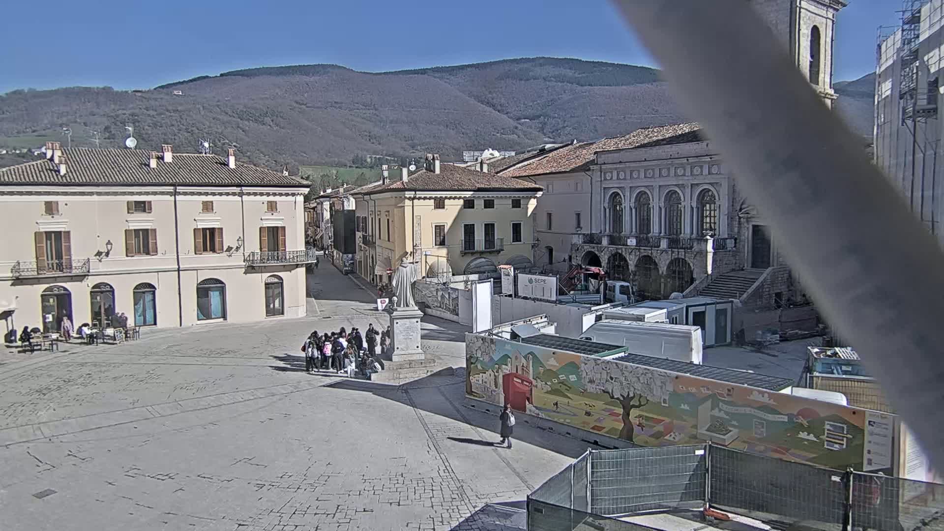 A town square with several buildings, a statue, and a group of people, under sunny skies and mountains in the background.