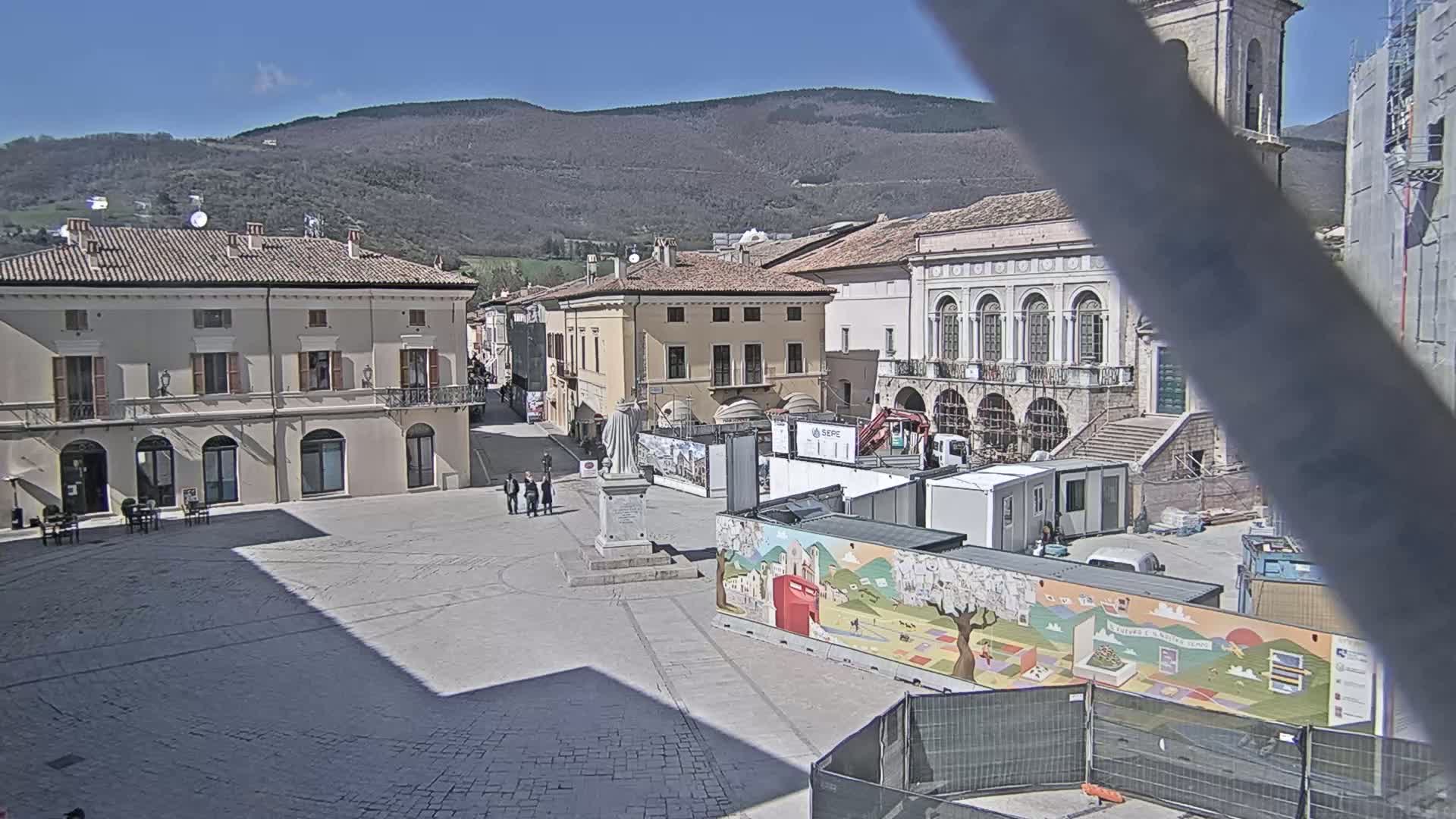 A town square under a clear sky, with several buildings surrounding it, some under construction, and a mountain in the background.