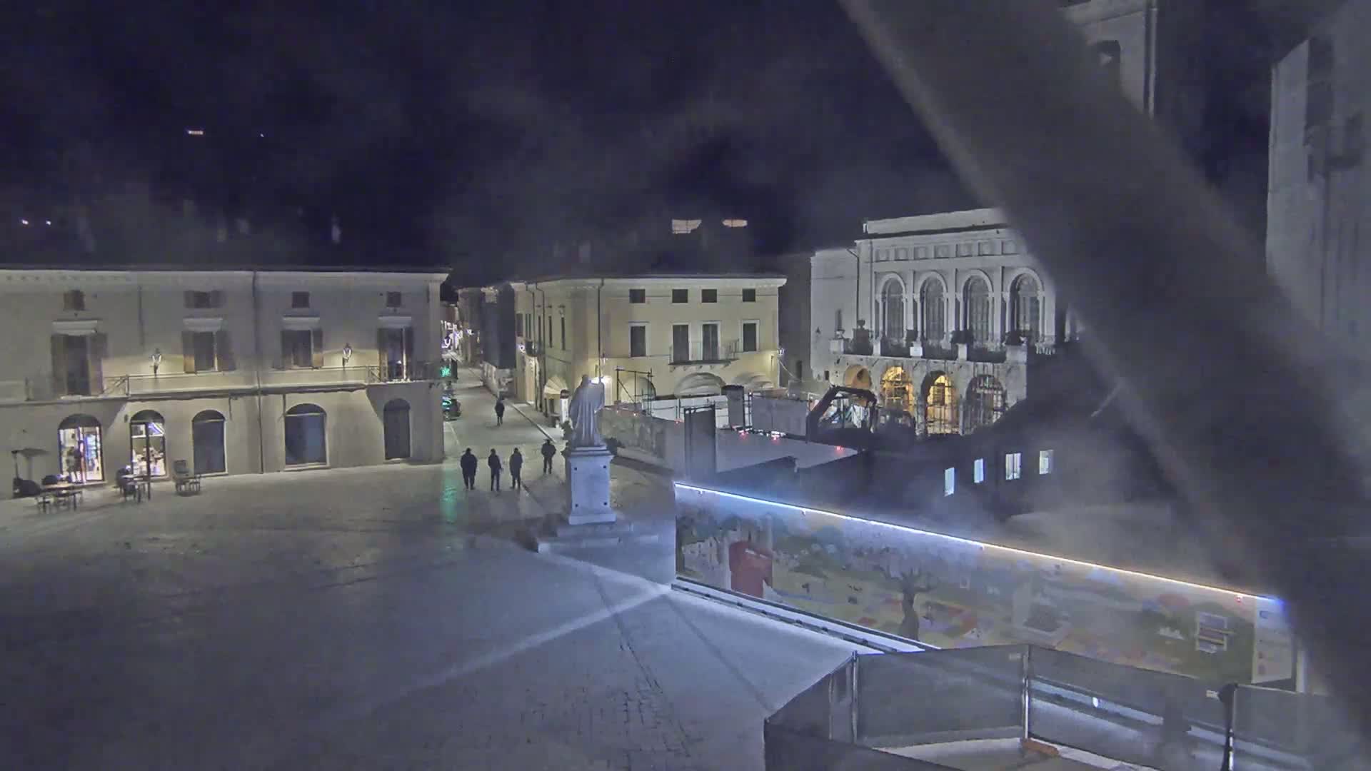 A nighttime view of a snow-covered town square with several buildings, a statue, and a few people walking.