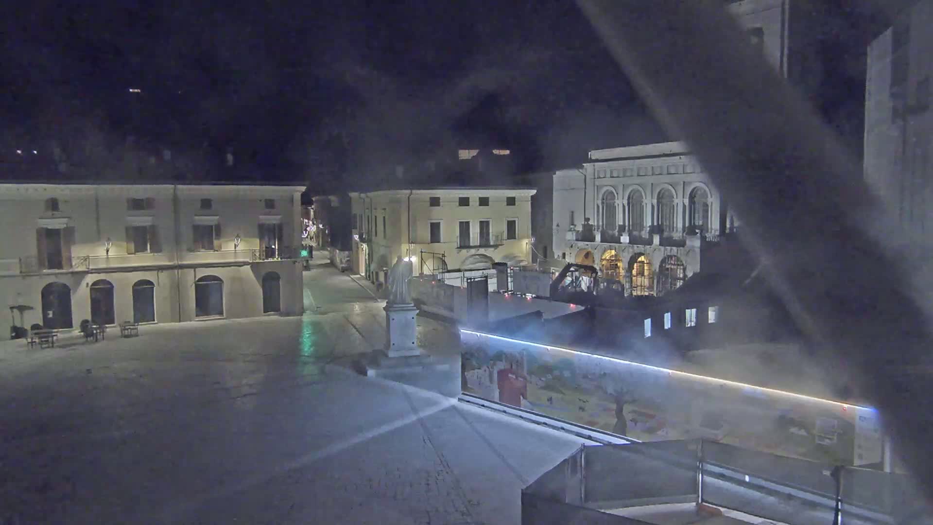 A nighttime view of a mostly empty town square with several buildings surrounding it, under hazy conditions.