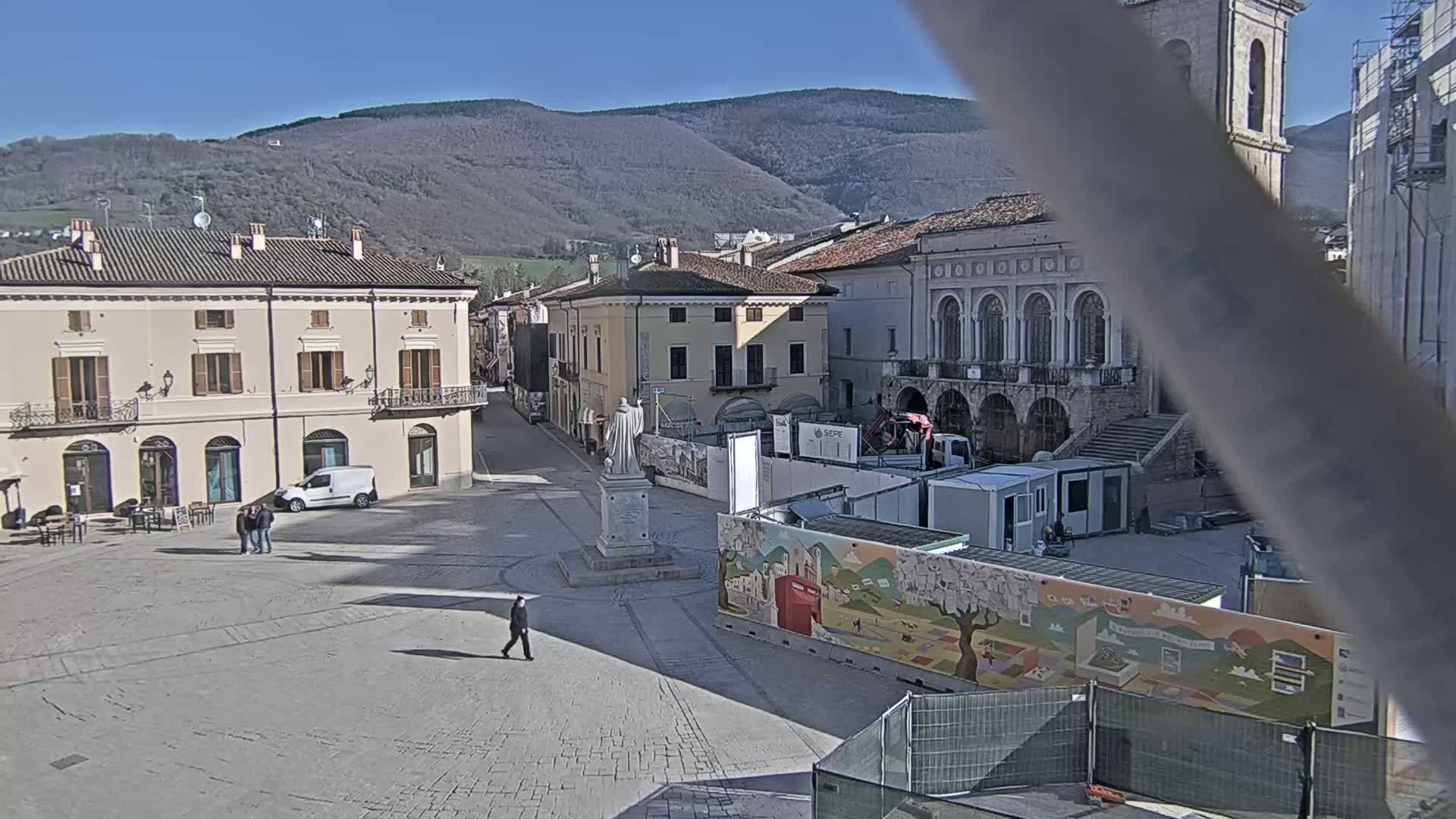 A town square, under sunny skies, features several buildings, a statue, construction barriers with colorful artwork, and a few pedestrians.