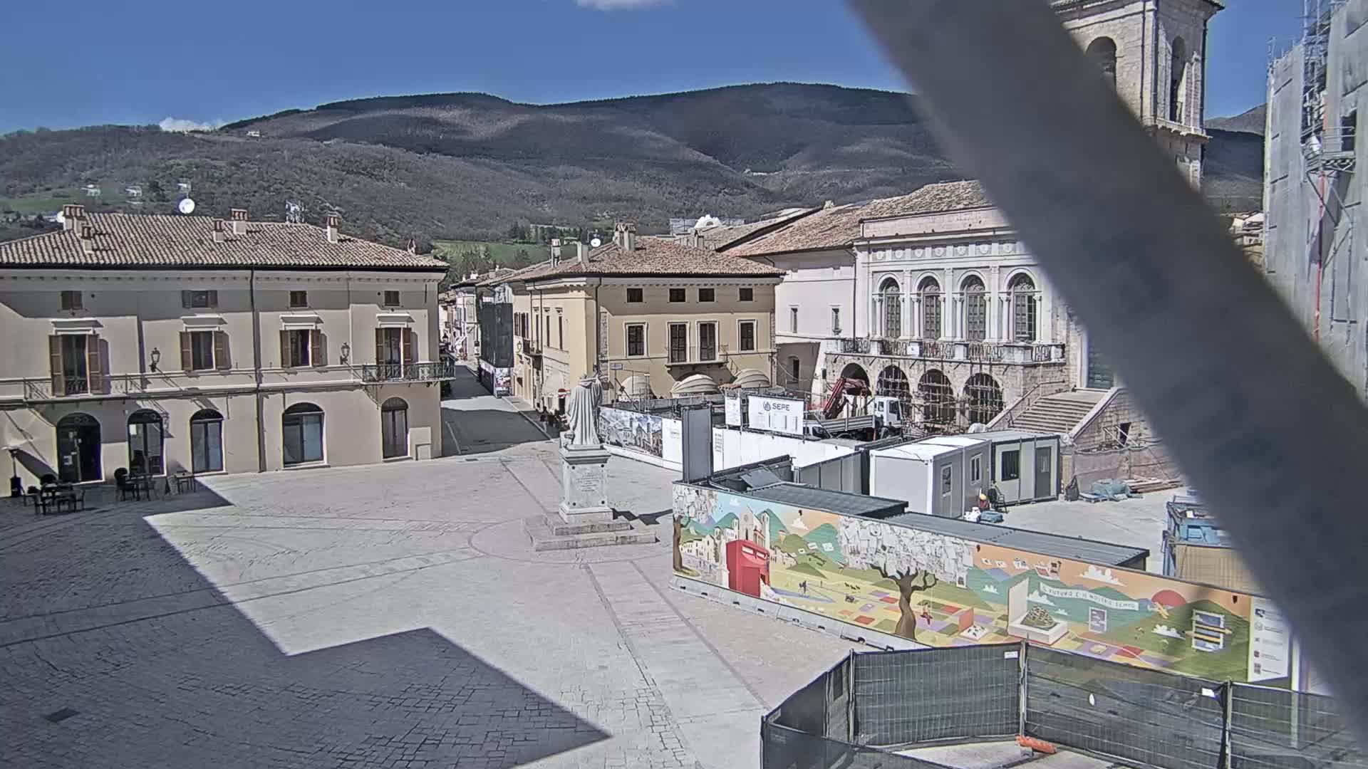 A town square in a sunny location, surrounded by historic buildings, with construction underway and a statue in the center, is set against a backdrop of mountains.