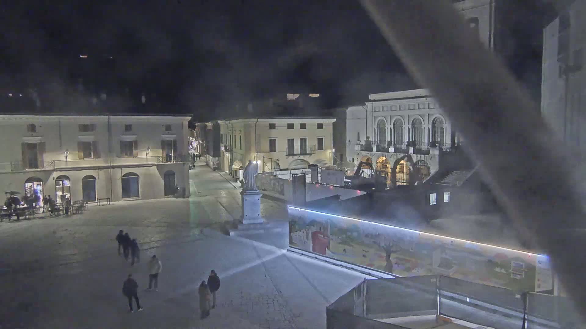 A nighttime view of a snow-covered town square with several people walking around, surrounded by buildings, one of which is under construction.