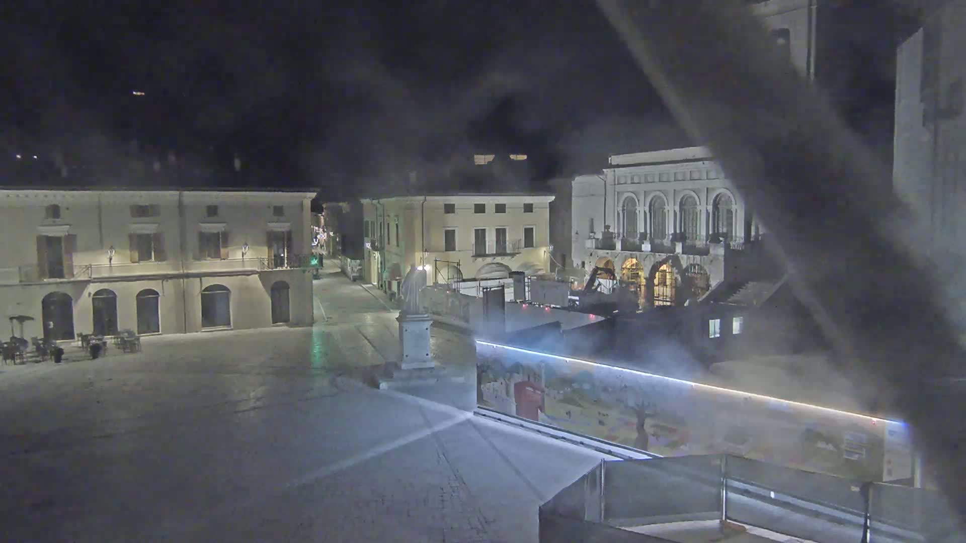A nighttime view of a town square with several buildings, a statue, and a construction barrier, under hazy or foggy conditions.