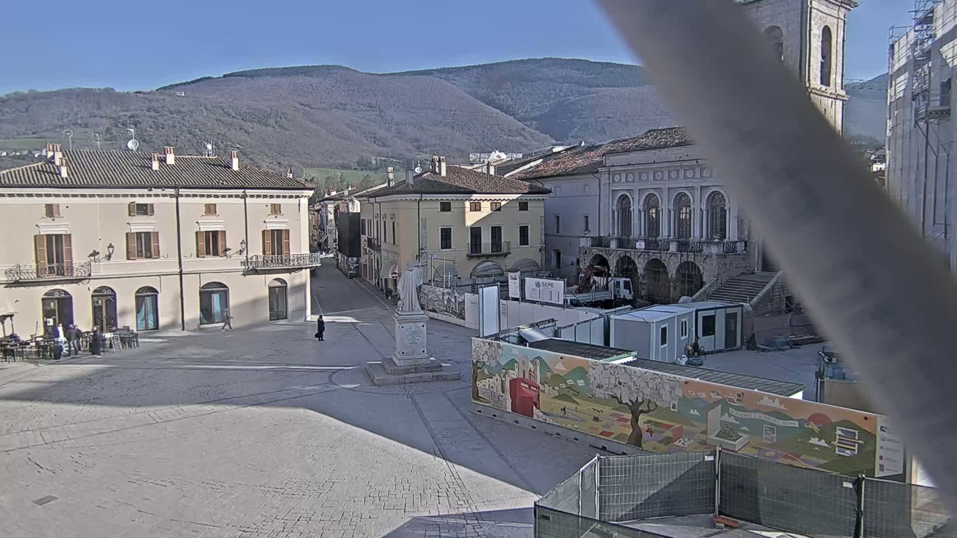 A sunny day reveals a town square under construction, with several buildings and mountains in the background.